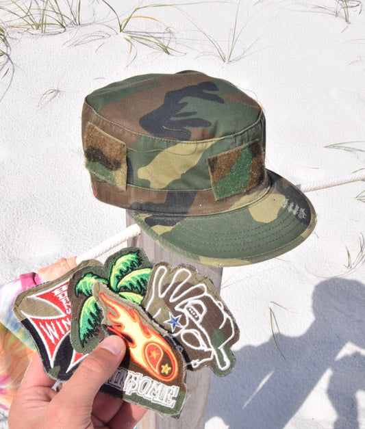 Camouflage hat held by a person with colorful stickers on a sandy background